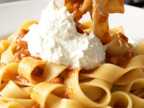 A close-up of a white plate filled with pappardelle pasta tossed in a thick, rich pork ragu with a dollop of ricotta.