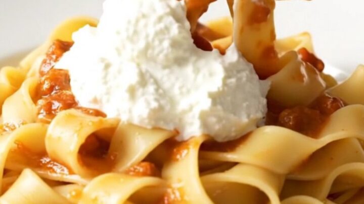A close-up of a white plate filled with pappardelle pasta tossed in a thick, rich pork ragu with a dollop of ricotta.