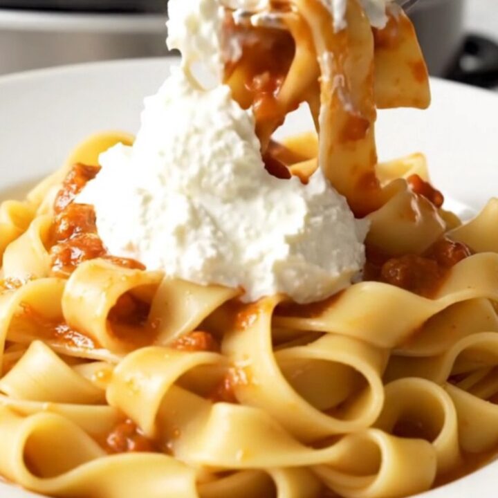 A close-up of a white plate filled with pappardelle pasta tossed in a thick, rich pork ragu with a dollop of ricotta.