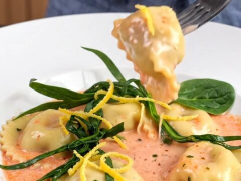A white bowl filled with tender lobster ravioli in a rich pink sauce, garnished with fresh tarragon and lemon zest.