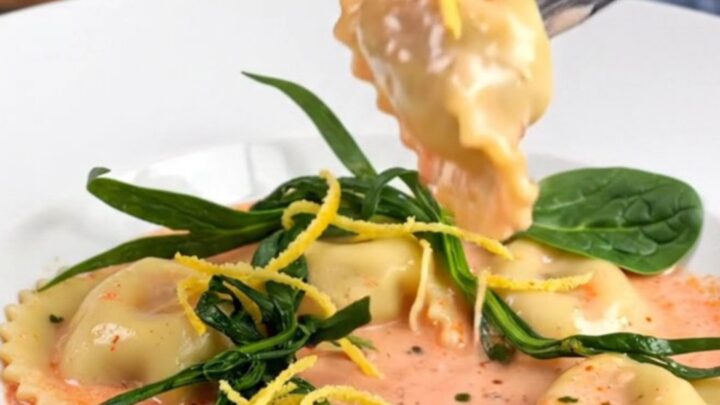 A white bowl filled with tender lobster ravioli in a rich pink sauce, garnished with fresh tarragon and lemon zest.