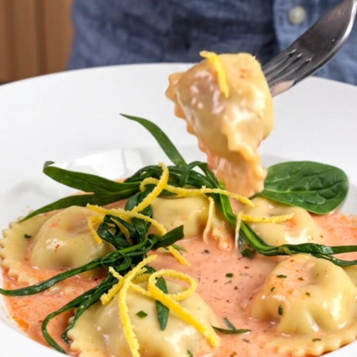 A white bowl filled with tender lobster ravioli in a rich pink sauce, garnished with fresh tarragon and lemon zest.