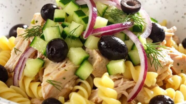 A close-up of creamy Greek chicken pasta in a turquoise slow cooker, topped with olives, cucumber, and fresh dill.