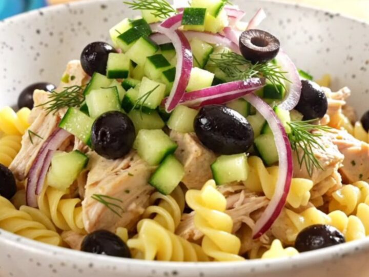 A close-up of creamy Greek chicken pasta in a turquoise slow cooker, topped with olives, cucumber, and fresh dill.