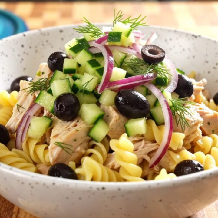 A close-up of creamy Greek chicken pasta in a turquoise slow cooker, topped with olives, cucumber, and fresh dill.