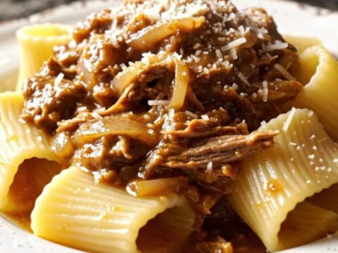 A close-up of paccheri pasta tubes coated in a rich, golden-brown beef and onion Genovese ragu on a dark plate.
