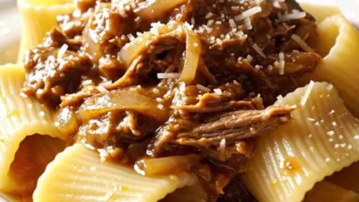 A close-up of paccheri pasta tubes coated in a rich, golden-brown beef and onion Genovese ragu on a dark plate.