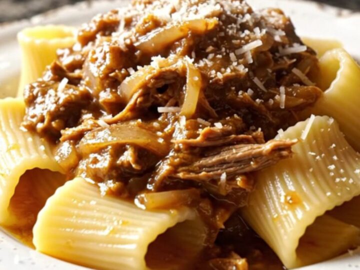 A close-up of paccheri pasta tubes coated in a rich, golden-brown beef and onion Genovese ragu on a dark plate.