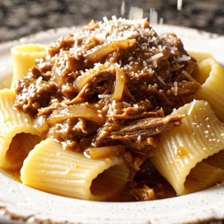 A close-up of paccheri pasta tubes coated in a rich, golden-brown beef and onion Genovese ragu on a dark plate.