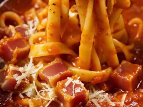 A close-up of bucatini pasta in a glossy tomato sauce with crispy guanciale pieces and grated pecorino cheese.