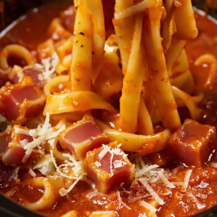 A close-up of bucatini pasta in a glossy tomato sauce with crispy guanciale pieces and grated pecorino cheese.