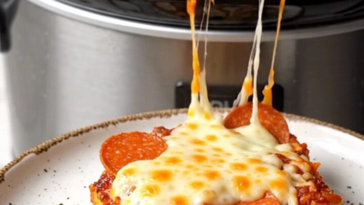 A close-up shot of a creamy pepperoni pizza pasta bake in a black crockpot with a long mozzarella cheese pull.