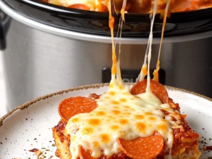 A close-up shot of a creamy pepperoni pizza pasta bake in a black crockpot with a long mozzarella cheese pull.