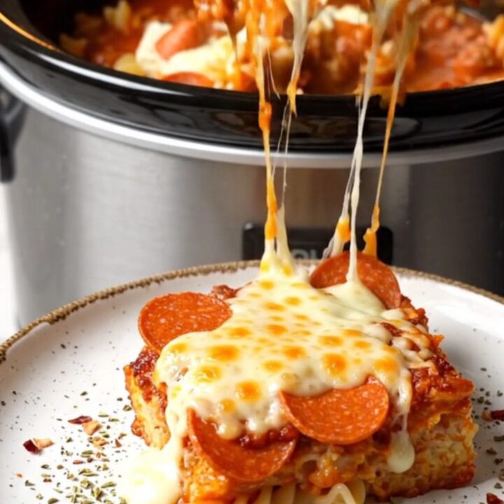 A close-up shot of a creamy pepperoni pizza pasta bake in a black crockpot with a long mozzarella cheese pull.