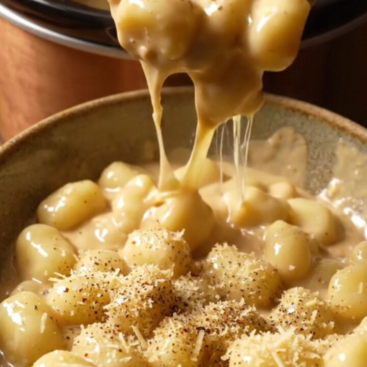 A close-up view of pillowy potato gnocchi covered in a thick, molten four-cheese cream sauce in a ceramic pot.