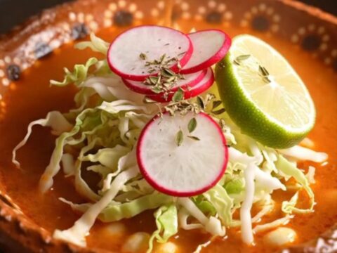 A steaming bowl of Posole Rojo topped with fresh radishes, cabbage, and lime, set in a cozy, warm-lit kitchen.