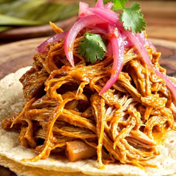 A steaming bowl of shredded Cochinita Pibil served on warm tortillas with pickled onions and cilantro.