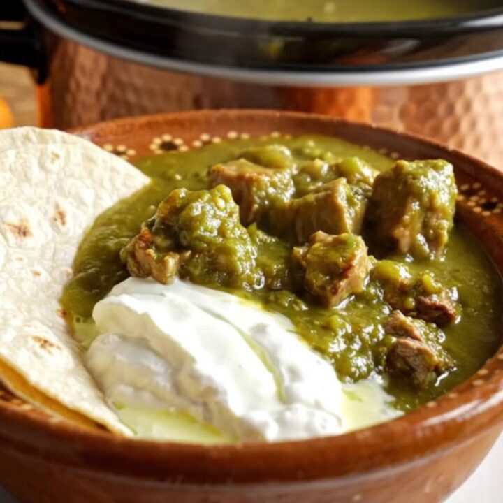 A rustic bowl of Green Chile Pork Chile Verde topped with crema and served with warm flour tortillas.