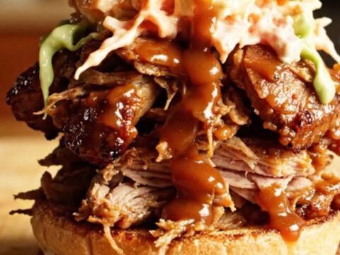 A close-up of a glossy, shredded root beer BBQ pork sandwich piled high on a toasted brioche bun with coleslaw.