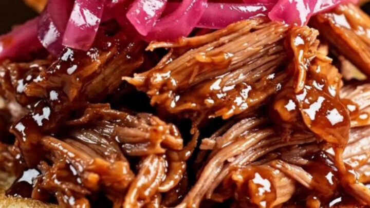 A glossy cherry-red slow cooker filled with tender, mahogany-colored pulled pork in a rich cherry glaze.