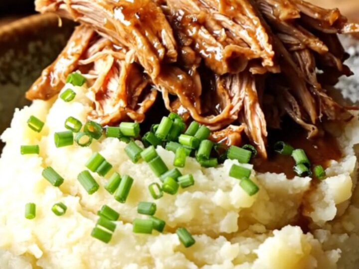 A close-up of tender pulled pork in a dark, rich gravy served over creamy mashed potatoes in a rustic kitchen.
