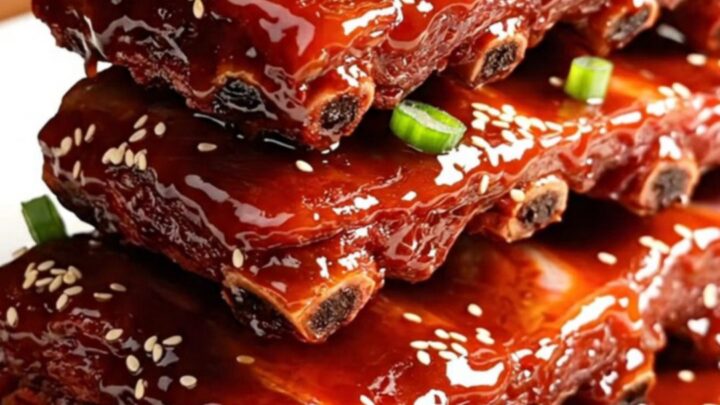 A glossy, dark red rack of Char Siu ribs stacked on a white ceramic platter, garnished with green onions and sesame seeds.