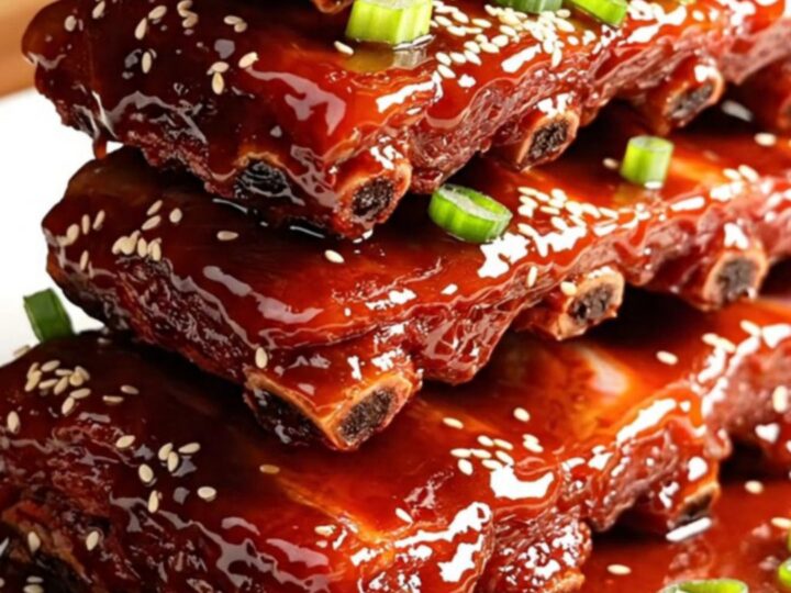 A glossy, dark red rack of Char Siu ribs stacked on a white ceramic platter, garnished with green onions and sesame seeds.