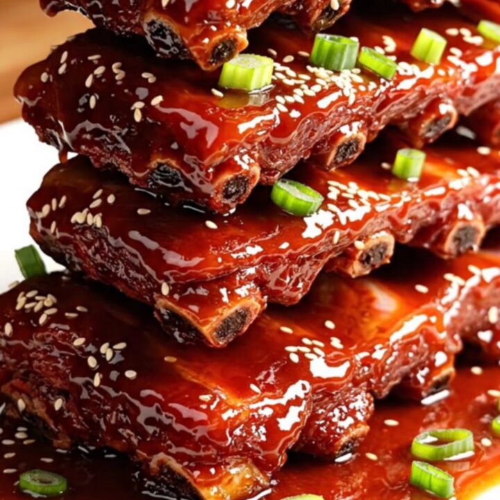 A glossy, dark red rack of Char Siu ribs stacked on a white ceramic platter, garnished with green onions and sesame seeds.
