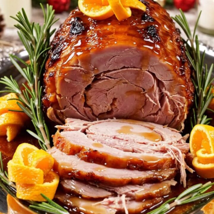 A festive spiral-cut ham glazed in a glossy amber sauce served on a silver platter with orange slices and rosemary.