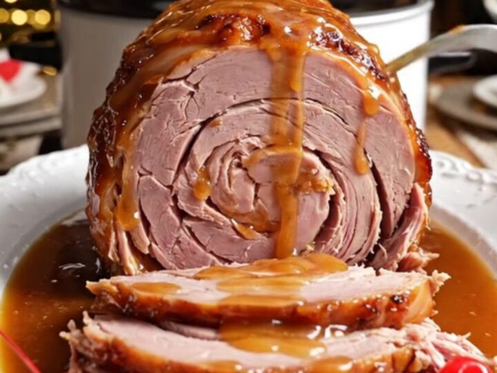 A glistening spiral-sliced ham in a white ceramic crockpot topped with pineapple rings and cherries.