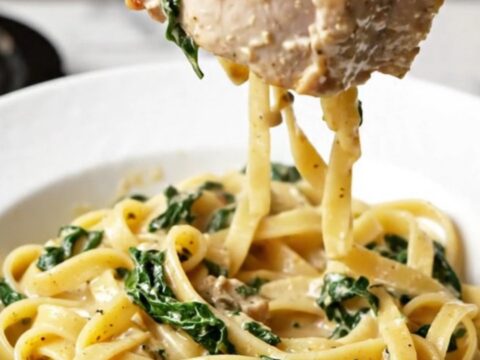 A plate of creamy Tuscan pork chops served over a bed of fettuccine with visible sun-dried tomatoes and spinach.