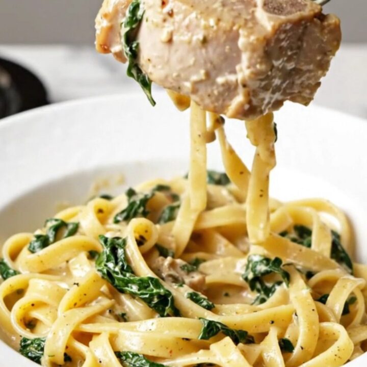A plate of creamy Tuscan pork chops served over a bed of fettuccine with visible sun-dried tomatoes and spinach.