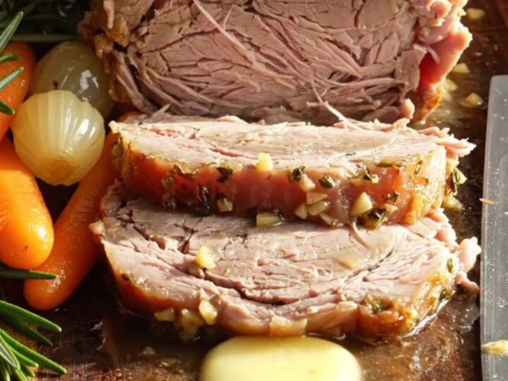A golden-brown sliced pork loin roast served on a wooden board with baby carrots and onions.