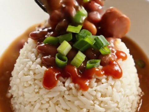 A close-up of a dark stoneware slow cooker filled with creamy red kidney beans, sliced Andouille sausage, and ham.