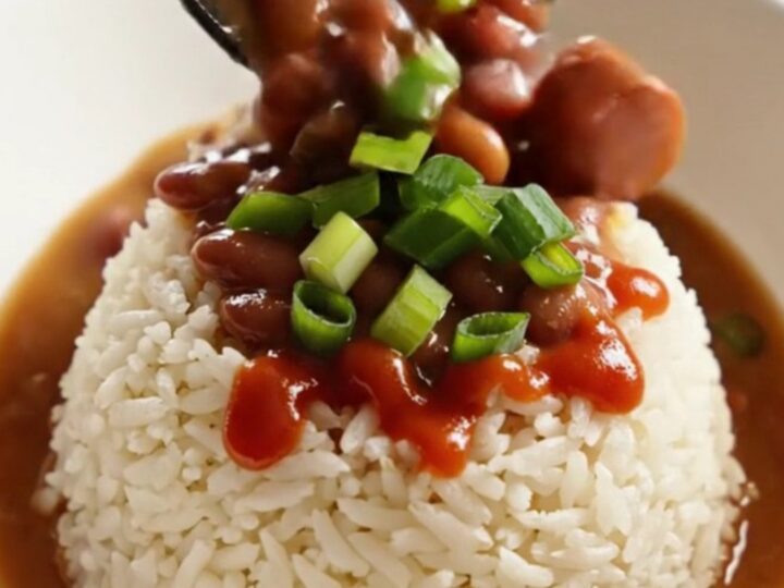 A close-up of a dark stoneware slow cooker filled with creamy red kidney beans, sliced Andouille sausage, and ham.