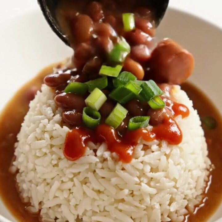 A close-up of a dark stoneware slow cooker filled with creamy red kidney beans, sliced Andouille sausage, and ham.