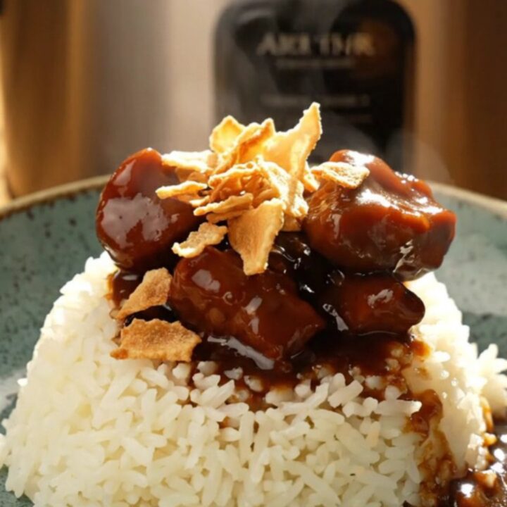 A bowl of glossy Filipino Pork Adobo served over steaming white jasmine rice, topped with golden crispy garlic chips.