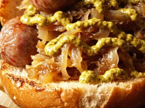 Plump, golden-brown bratwurst links nestled in a bed of sauerkraut and caramelized apples in a rustic stoneware slow cooker.