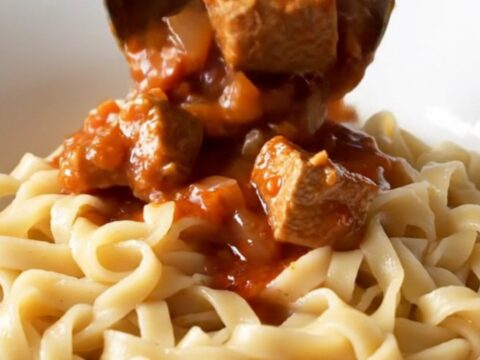 A bowl of warm, creamy pork goulash with tender meat and a rich red paprika sauce over egg noodles.