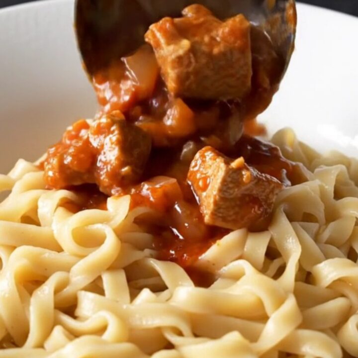 A bowl of warm, creamy pork goulash with tender meat and a rich red paprika sauce over egg noodles.