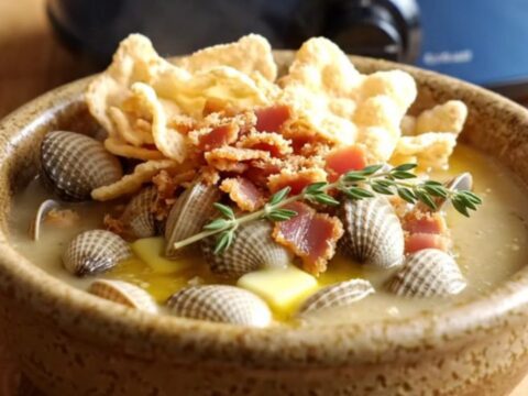 A steaming bowl of creamy New England clam and corn chowder topped with oyster crackers, bacon, and fresh thyme.