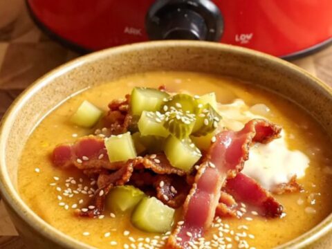 A close-up of a cherry-red ceramic Crockpot filled with rich, golden cheeseburger chowder with a large ladle lifting