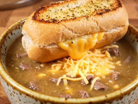 A warm ceramic bowl filled with creamy Philly cheesesteak soup topped with a toasted garlic bread slice.
