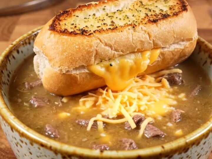 A warm ceramic bowl filled with creamy Philly cheesesteak soup topped with a toasted garlic bread slice.