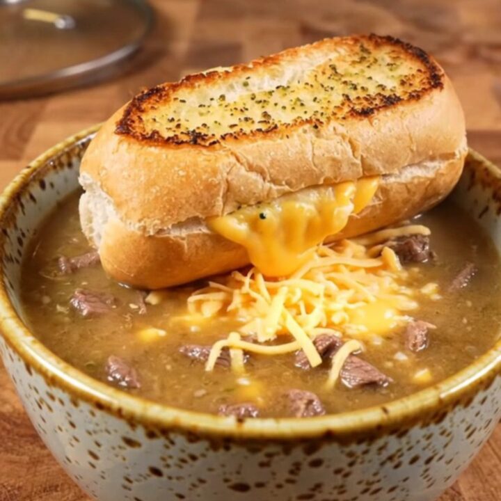 A warm ceramic bowl filled with creamy Philly cheesesteak soup topped with a toasted garlic bread slice.