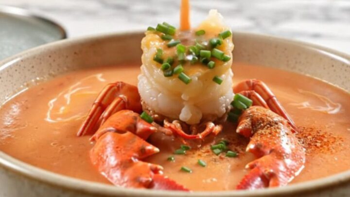 A creamy coral-pink lobster bisque in a white porcelain bowl, topped with a lobster claw and chives.