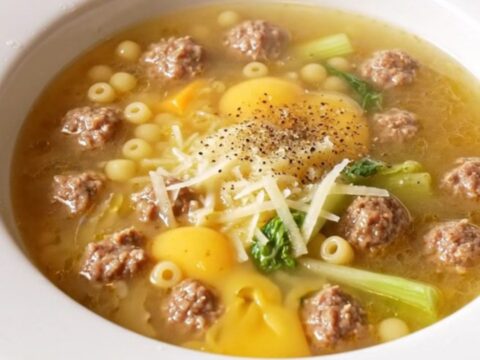 A wide rimmed bowl filled with Italian wedding soup featuring mini meatballs, pearl pasta, and escarole.