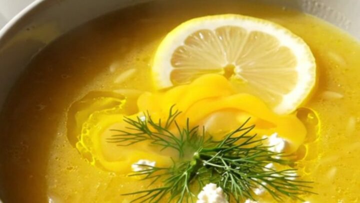 A steaming bowl of creamy Greek lemon chicken orzo soup garnished with fresh dill, feta, and a lemon slice.