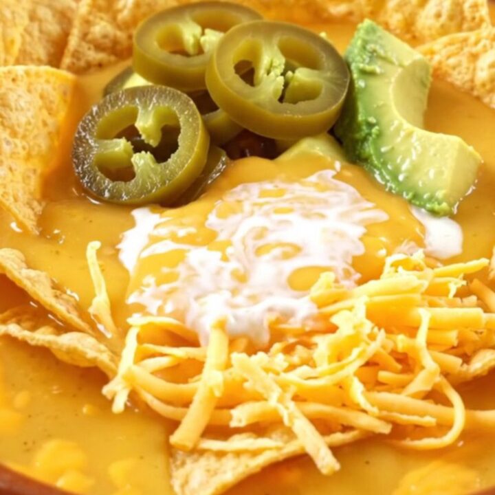 A warm ceramic crockpot filled with creamy nacho beef soup, topped with tortilla chips, avocado, and pickled jalapeños.