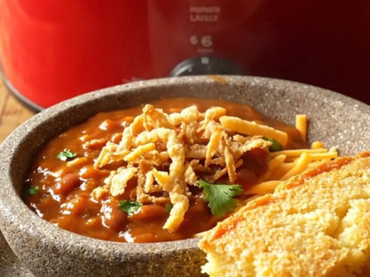 A close-up shot of a deep red ceramic slow cooker filled with glossy, sweet-heat BBQ chicken chili with melted cheese.